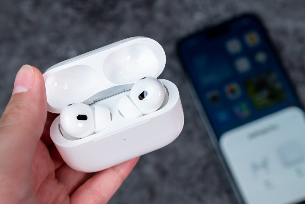 Apple AirPods In the woman's hand, there is an open case with headphones—AirPods Pro 2 generation. In the background, there is an out-of-focus smartphone that is connected to AirPods.